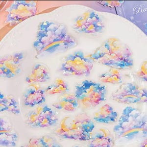 May include: A selection of cloud-shaped stickers in pastel shades of pink, blue, yellow, and purple, with rainbows, moons, and stars. The stickers are on a white surface. The word "Rainbow" is visible on the packaging.