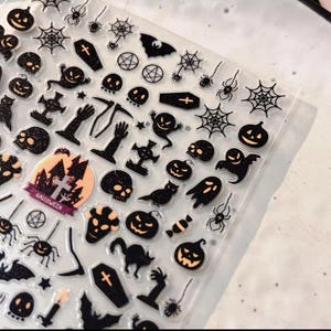 May include: A sheet of Halloween-themed stickers featuring various spooky designs. Includes black spiders, cobwebs, ghosts, jack-o'-lanterns, skulls, bats, and coffins. The stickers have a glittery finish and are arranged on a clear backing.
