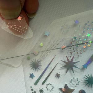 May include: A clear nail with a star design is held above a sheet of iridescent star and comet nail art stickers. The stickers feature various star shapes and comet trails in holographic and metallic finishes, perfect for nail art.