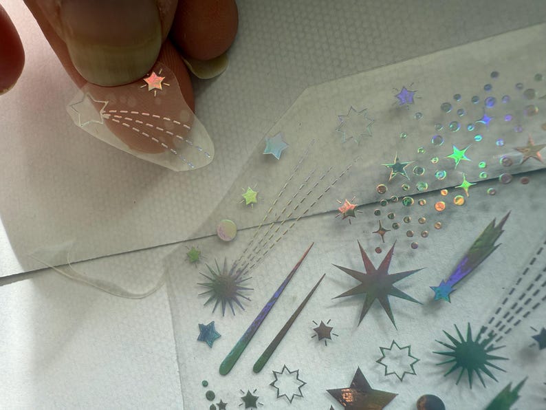 May include: A clear nail with a star design is held above a sheet of iridescent star and comet nail art stickers. The stickers feature various star shapes and comet trails in holographic and metallic finishes, perfect for nail art.