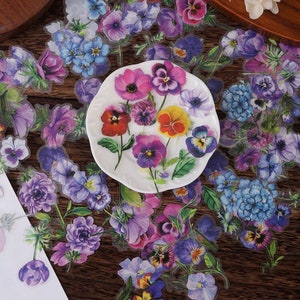 May include: A sheet of clear stickers featuring various colorful floral designs. The stickers feature purple, pink, blue, and yellow flowers with green leaves. The stickers are arranged on a wooden surface.
