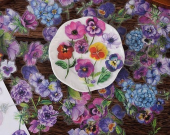 May include: A sheet of clear stickers featuring various colorful floral designs. The stickers feature purple, pink, blue, and yellow flowers with green leaves. The stickers are arranged on a wooden surface.