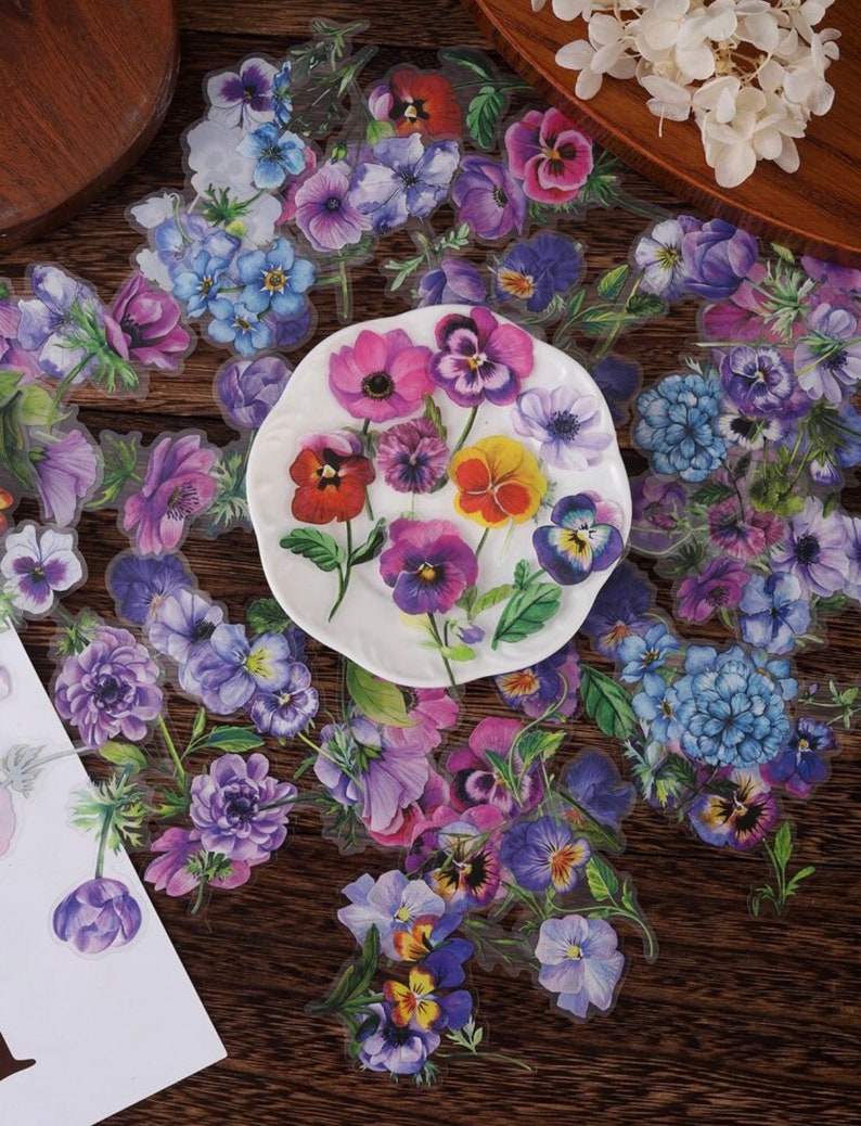 May include: A sheet of clear stickers featuring various colorful floral designs. The stickers feature purple, pink, blue, and yellow flowers with green leaves. The stickers are arranged on a wooden surface.