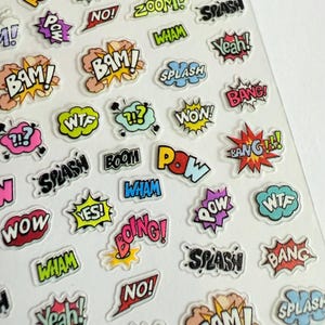 May include: Sheet of comic book-style stickers with various action words and sound effects. The stickers feature words like "POW," "BAM," "SPLASH," and "WOW" in colorful speech bubbles and starburst shapes. Ideal for scrapbooking or adding flair to crafts.