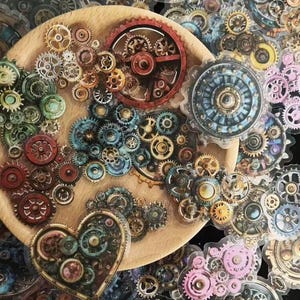 Könnte beinhalten: Eine Sammlung von Aufklebern mit Zahnrädern im Steampunk-Stil in verschiedenen Größen und Farben, darunter Gold, Bronze, Rot, Blau und Rosa. Einige Zahnräder sind auf einem Holzbrett angeordnet, andere sind verstreut.