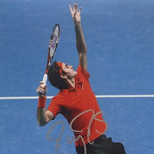 May include: A tennis player in a red shirt and headband serves a tennis ball. The player is holding a tennis racket with a white handle and a red and black head. The background is a solid blue color.