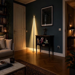 Puede incluir: Una l&aacute;mpara de pie negra moderna con un dise&ntilde;o vertical, que emite una luz c&aacute;lida y enfocada. La l&aacute;mpara se encuentra en una habitaci&oacute;n con paredes de color verde azulado oscuro, una alfombra estampada y un mueble decorativo. El ambiente es acogedor.