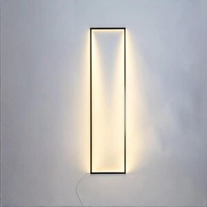 Front view of warm-lit minimalist floor lamp in modern living space. Sleek black frame casts soft glow on neutral walls. Perfect for cozy evenings.