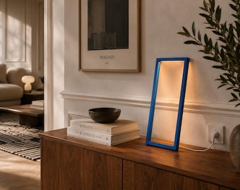 May include: A modern blue rectangular LED lamp with a warm glow, resting on a dark wood cabinet. The lamp is angled, showcasing its illuminated interior. A stack of books and a dark bowl are also on the cabinet. A framed print hangs on the wall.