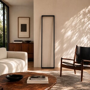 Op de afbeelding: Een moderne woonkamer met een lichtgekleurde bank, een houten salontafel en een stoel met zwart frame. Een groot zwart rechthoekig frame staat tegen een neutrale muur. Een houten kast en ingelijste kunstwerken zijn zichtbaar.