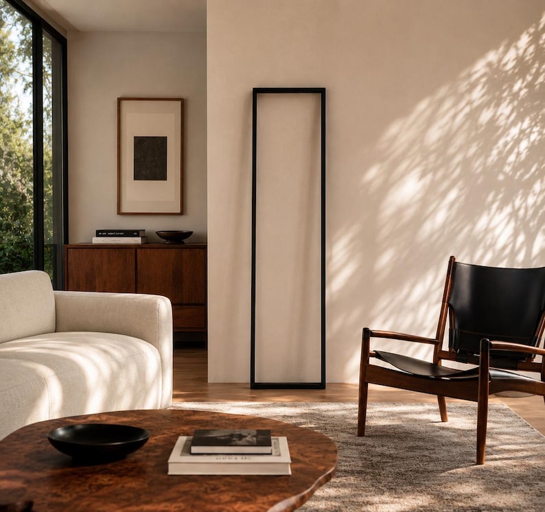 Op de afbeelding: Een moderne woonkamer met een lichtgekleurde bank, een houten salontafel en een stoel met zwart frame. Een groot zwart rechthoekig frame staat tegen een neutrale muur. Een houten kast en ingelijste kunstwerken zijn zichtbaar.