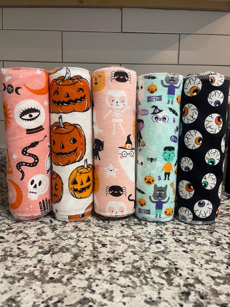 May include: Five cylindrical rolls of fabric featuring Halloween-themed patterns. The prints include orange pumpkins, skulls, snakes, ghosts, spiders, skeletons, monsters, and eyeballs on backgrounds of pink, white, light blue, and black.