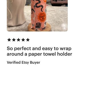 May include: A cylindrical fabric roll featuring a repeating pattern of black and orange celestial motifs, including suns, moons, crystal balls, and hands on a light pink background, secured with visible stitching along the vertical edge.