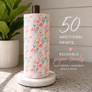 Può includere: Un rotolo di carta assorbente riutilizzabile con motivo floreale rosa, arancione e verde acqua su sfondo bianco, montato su una base circolare bianca. Il testo recita: 50 ADDITIONAL PRINTS. REUSABLE paper towels ECO-FRIENDLY. ABSORBENT. WASH & REUSE.