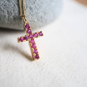 May include: A gold-colored cross pendant necklace with a chain. The cross is encrusted with small, bright pink gemstones. The necklace is displayed on a white surface with a gray stone in the background.