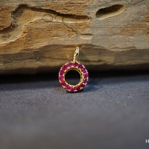 May include: A gold-colored pendant with a circular design, encrusted with numerous small, vibrant red gemstones. The pendant is set against a dark gray surface with a wooden background, creating a contrast that highlights the jewelry's details.