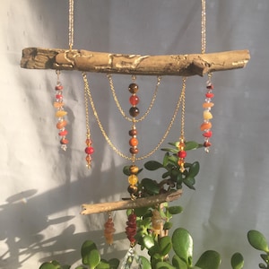 Op de afbeelding: Een handgemaakte windgong met een natuurlijke houten tak versierd met goudkleurige kettingen en strengen kleurrijke edelstenen. De edelstenen omvatten rode, oranje, gele en bruine kralen. Een helder kristallen prisma hangt aan de onderkant.