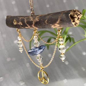Op de afbeelding: Een decoratief hangornament met een donkerbruine houten tak. Gouden kettingen hangen heldere kristallen kralen, een blauwe steen en een gouden vredesteken op. Het ornament is ontworpen voor woondecoratie.