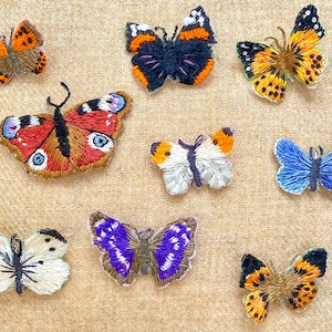 Könnte beinhalten: Eine Sammlung von acht gestickten Schmetterlings-Patches in verschiedenen Farben und Mustern, darunter Orange, Schwarz, Weiß, Blau, Rot und Lila.