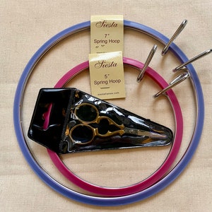 Könnte beinhalten: Zwei Stickrahmen, einer mit 12,7 cm und einer mit 17,8 cm Durchmesser, beide in einem Lila- und Rosaton. Eine goldene Stickerschere ist in einer durchsichtigen Plastiktüte enthalten.