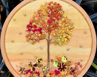 May include: Embroidered hoop art featuring a stylized tree with a colorful autumn foliage of red, orange, yellow, and green beads. The tree is surrounded by a variety of colorful beads and sequins.