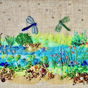 Peut inclure: Un gros plan sur une broderie au point d'aiguille représentant une scène fantaisiste avec deux libellules bleues, une végétation verte et un cours d'eau bleu. La broderie est réalisée sur un fond de lin beige et présente une variété de perles et de paillettes colorées.