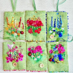 Könnte beinhalten: Sechs grüne Filz-Anhänger mit gestickten Blumenmotiven. Jeder Anhänger zeigt eine andere Blumenart, darunter rote Mohnblumen, Glockenblumen und rosa Blüten. Die Anhänger sind mit Pailletten und Perlen verziert.