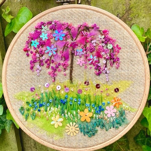 May include: Embroidered hoop art featuring a pink and purple weeping willow tree with a green and blue grass base. The tree is decorated with sequins and beads.