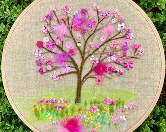 May include: Embroidered hoop art featuring a pink and white tree with a brown trunk and green leaves. The tree is surrounded by a field of pink, white, and green flowers.