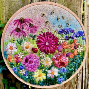 Pode incluir: Um design floral bordado redondo com uma variedade de flores coloridas, incluindo echinaceas rosa, margaridas e flores vibrantes. A obra de arte inclui grama verde, joaninhas e abelhas, tudo dentro de um círculo de madeira.