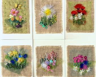 May include: A set of six handmade greeting cards featuring embroidered floral designs. Each card showcases a unique arrangement of colorful flowers, sequins, and beads on a burlap-textured background. The cards are mounted on white cardstock, creating a rustic and artistic aesthetic.