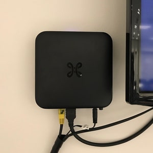 Op de afbeelding: Een zwart streamingapparaat met een logo aan de voorkant is aangesloten op een zwarte televisie met een blauw scherm. Het apparaat is met de televisie verbonden via zwarte kabels.