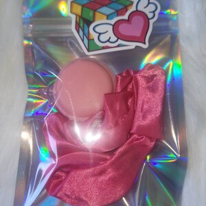 May include: A pink satin scrunchie and a pink round container in a clear iridescent bag with a sticker of a colorful Rubik's cube and a pink heart with wings.