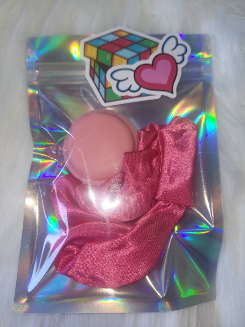 May include: A pink satin scrunchie and a pink round container in a clear iridescent bag with a sticker of a colorful Rubik's cube and a pink heart with wings.