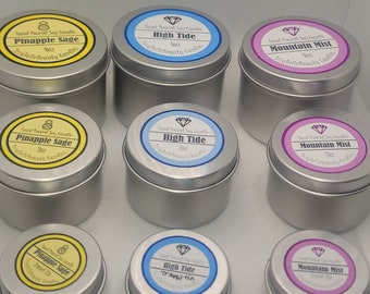 May include: Nine silver candle tins with labels. The labels are yellow, blue, and purple. The labels have the text "Hand Poured Soy Candle" and the names of the scents: "Pineapple Sage", "High Tide", and "Mountain Mist". The candle tins are 4 oz and 2 oz.