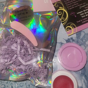 May include: A small pink lip balm jar with a red lip balm inside. The jar is in a clear plastic bag with purple shredded paper. The bag has a thank you sticker on it. The sticker says "Thank you for supporting my small business". There is a pink business card with the text "IzzaBelle Beauty Handmade beauty products. Natural, Organic, Cruelty free beauty products. Upgrade your beauty care. Jennifer Co Owner Instagram: @izzabe Twitter: @izzab Tiktok: @izzab Email for Custom orders: izzabellebs"