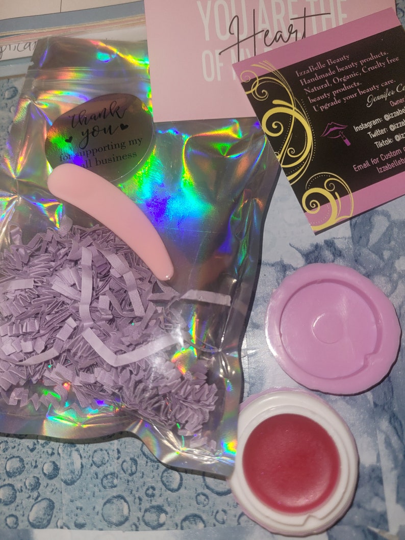 May include: A small pink lip balm jar with a red lip balm inside. The jar is in a clear plastic bag with purple shredded paper. The bag has a thank you sticker on it. The sticker says "Thank you for supporting my small business". There is a pink business card with the text "IzzaBelle Beauty Handmade beauty products. Natural, Organic, Cruelty free beauty products. Upgrade your beauty care. Jennifer Co Owner Instagram: @izzabe Twitter: @izzab Tiktok: @izzab Email for Custom orders: izzabellebs"