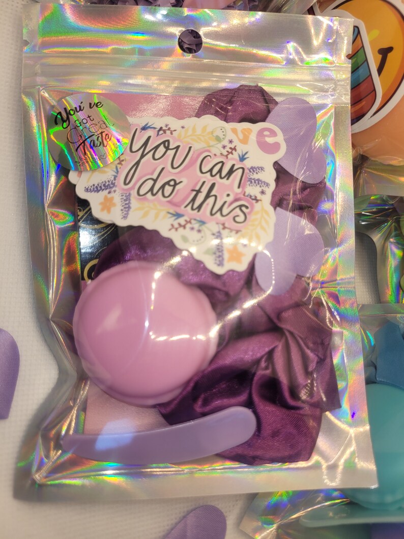 May include: A clear plastic bag filled with purple and pink items. The bag contains a purple satin scrunchie, a pink round compact, a purple nail file, and a sticker that says "You've got great taste" and another sticker that says "You can do this".