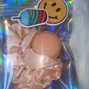 May include: A pink satin scrunchie and a peach-colored hair scrunchie in a clear iridescent bag with a smiley face sticker and a cartoon drawing of a rainbow slushie.