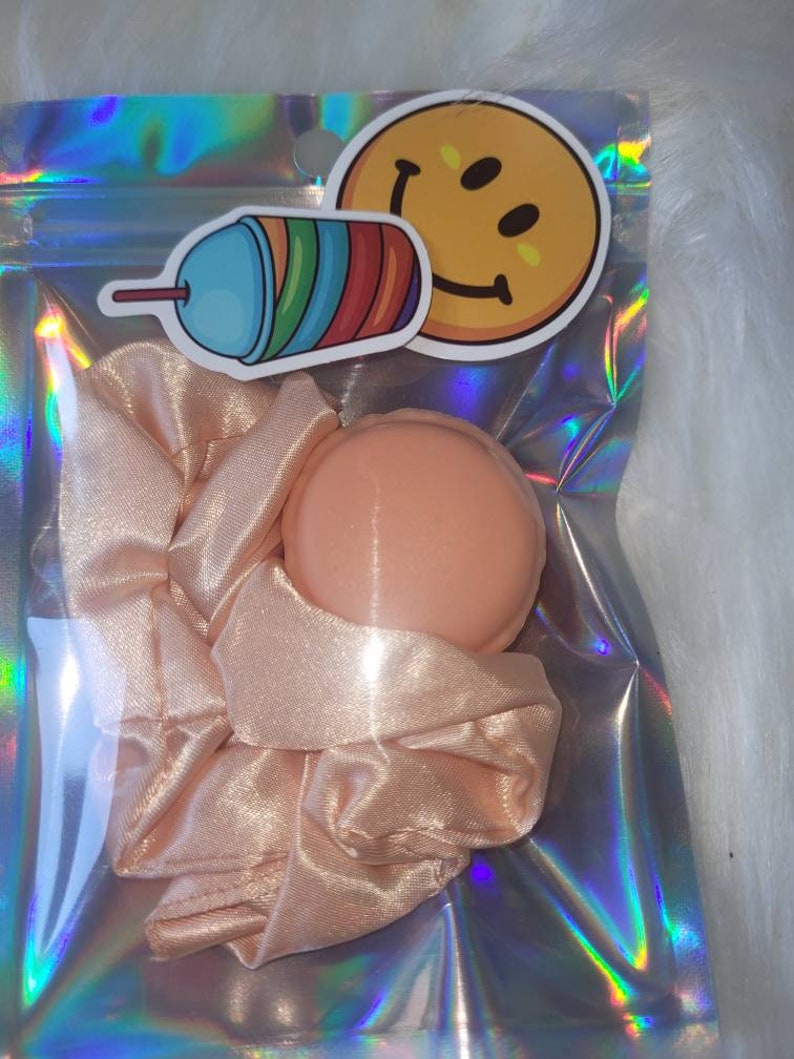 May include: A pink satin scrunchie and a peach-colored hair scrunchie in a clear iridescent bag with a smiley face sticker and a cartoon drawing of a rainbow slushie.