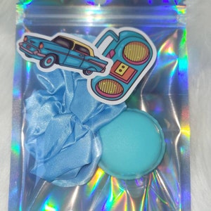 May include: A blue iridescent zippered pouch with a blue satin scrunchie, a blue round makeup sponge, and two stickers. One sticker is of a blue car and the other is of a retro boombox.