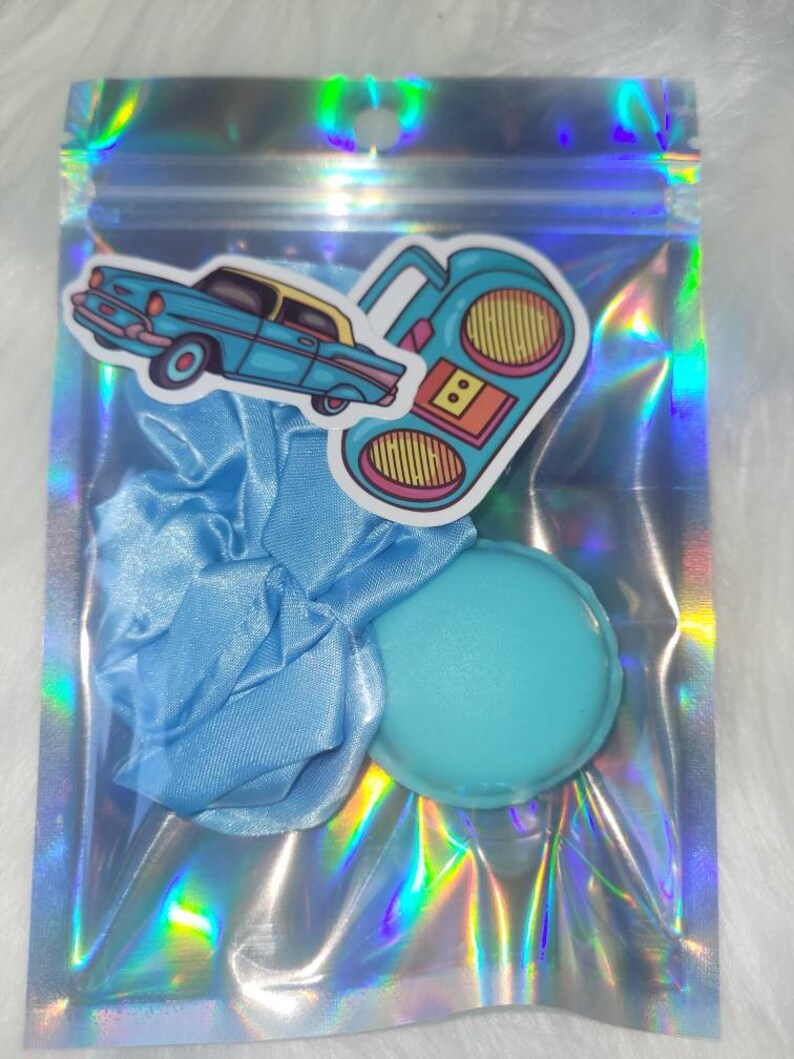 May include: A blue iridescent zippered pouch with a blue satin scrunchie, a blue round makeup sponge, and two stickers. One sticker is of a blue car and the other is of a retro boombox.