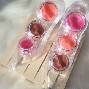 Puede incluir: Un conjunto de seis brillos de labios transparentes en un recipiente de plástico transparente con forma de zapato. Los brillos de labios son en tonos de rosa, rojo y naranja.