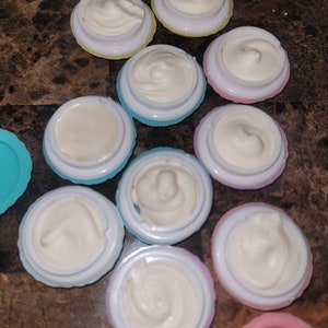 May include: A collection of 11 small, round containers with lids. The containers are filled with a white, creamy substance. The containers are in various colors, including blue, pink, yellow, and purple.