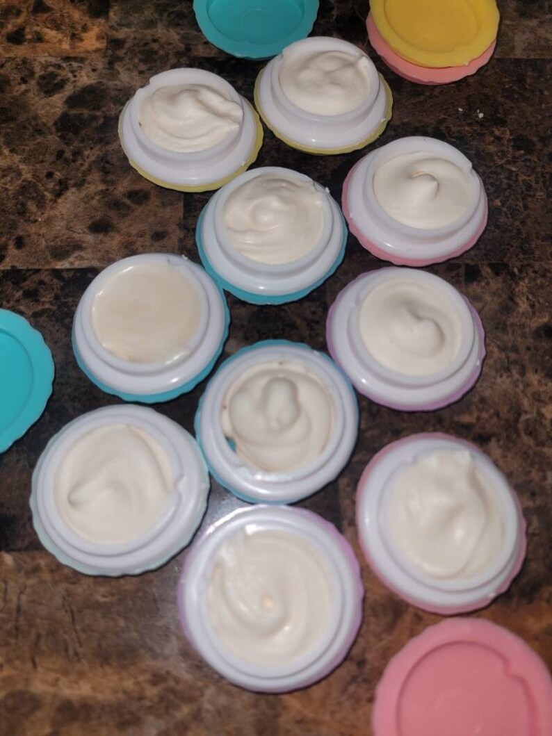May include: A collection of 11 small, round containers with lids. The containers are filled with a white, creamy substance. The containers are in various colors, including blue, pink, yellow, and purple.