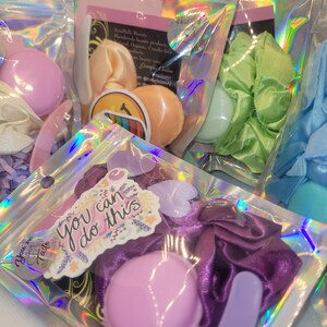May include: A set of three small, pastel-colored, round, decorative items, a purple satin scrunchie, a purple plastic spatula, and a sticker with the text "You can do this" in a floral design. The items are packaged in a clear plastic bag with a holographic iridescent pattern.
