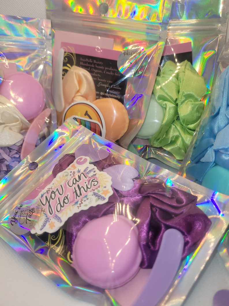 May include: A set of three small, pastel-colored, round, decorative items, a purple satin scrunchie, a purple plastic spatula, and a sticker with the text "You can do this" in a floral design. The items are packaged in a clear plastic bag with a holographic iridescent pattern.