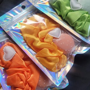 May include: Three iridescent zippered bags with satin scrunchies inside. The scrunchies are in green, yellow, and orange. Each bag has a white heart-shaped tag.
