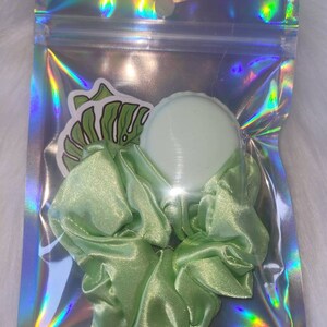 May include: A green satin scrunchie with a green plastic hair tie in a clear plastic bag with a holographic iridescent finish.