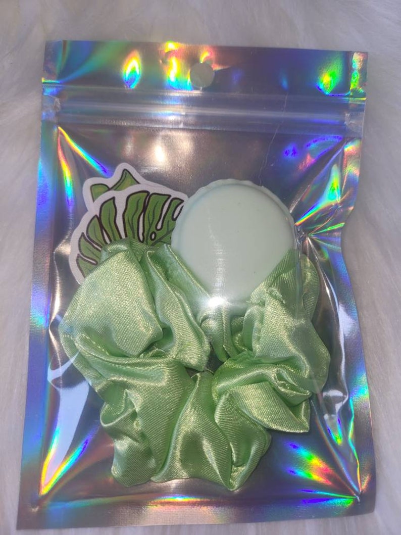 May include: A green satin scrunchie with a green plastic hair tie in a clear plastic bag with a holographic iridescent finish.